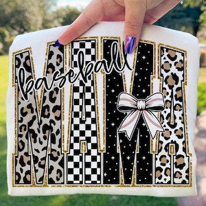 May include: White t-shirt featuring the word "MAMA" in large, decorative letters with a gold glitter outline. The letters are filled with leopard print, a black and white checkered pattern, and a black background with white stars. The word "baseball" is written in cursive.