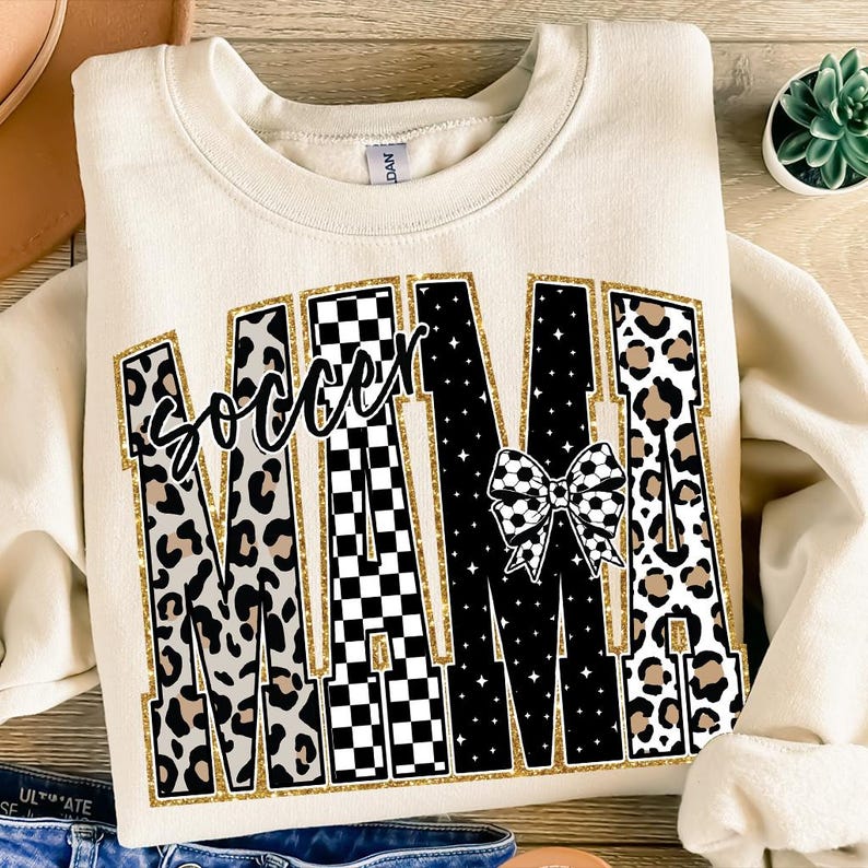 May include: Cream-colored sweatshirt with the text "Soccer Mom" in a bold, decorative font. The letters feature leopard print, black and white checkered patterns, and a soccer ball bow. The text is outlined in gold glitter.