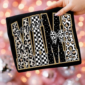 May include: Black t-shirt with the word "MAMA" in large, stylized letters. The letters feature leopard print, checkered patterns, and a soccer ball bow. The word "Soccer" is written above the "MAMA" text.