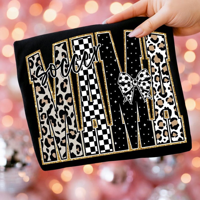 May include: Black t-shirt with the word "MAMA" in large, stylized letters. The letters feature leopard print, checkered patterns, and a soccer ball bow. The word "Soccer" is written above the "MAMA" text.