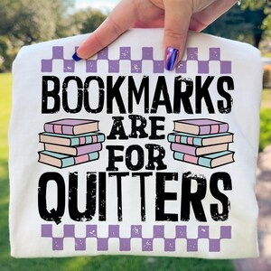 May include: White t-shirt with the text "BOOKMARKS ARE FOR QUITTERS" in black, with stacks of books and a purple checkered border. The shirt is held up by a hand.