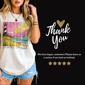 May include: Cream-colored t-shirt featuring a colorful beach scene with a lighthouse, ocean, and text that reads "Be salty. Stay lit." The shirt also has the text "Thank You" on a black background.