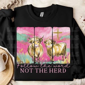 Op de afbeelding: Zwart sweatshirt met een afbeelding van twee Schotse Hooglandrunderen tegen een roze en groene achtergrond. De tekst "Follow the word NOT THE HERD" en "ISAIAH 8:11-13" is eronder gedrukt. Het sweatshirt is gemaakt van zacht materiaal.
