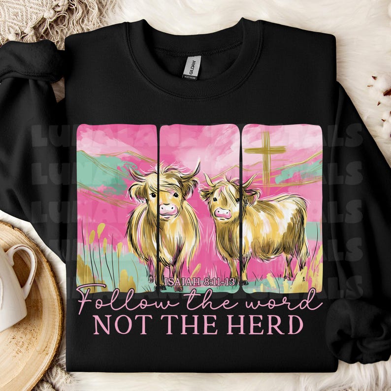 Op de afbeelding: Zwart sweatshirt met een afbeelding van twee Schotse Hooglandrunderen tegen een roze en groene achtergrond. De tekst "Follow the word NOT THE HERD" en "ISAIAH 8:11-13" is eronder gedrukt. Het sweatshirt is gemaakt van zacht materiaal.