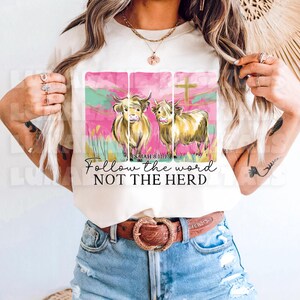 Op de afbeelding: Cr&egrave;mekleurig T-shirt met een afbeelding van twee Schotse Hooglandrunderen, een kruis en de tekst "Follow the word NOT THE HERD". Het ontwerp bevat roze, groene en gouden accenten.