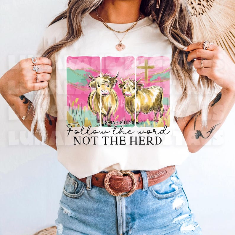 Op de afbeelding: Cr&egrave;mekleurig T-shirt met een afbeelding van twee Schotse Hooglandrunderen, een kruis en de tekst "Follow the word NOT THE HERD". Het ontwerp bevat roze, groene en gouden accenten.