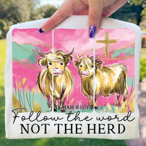 Op de afbeelding: Wit T-shirt met een kleurrijke illustratie van twee Highland-koeien tegen een roze en groene achtergrond, met een gouden kruis en de tekst "Follow the word NOT THE HERD". De tekst "Jesaja 8:11-13" is ook inbegrepen.