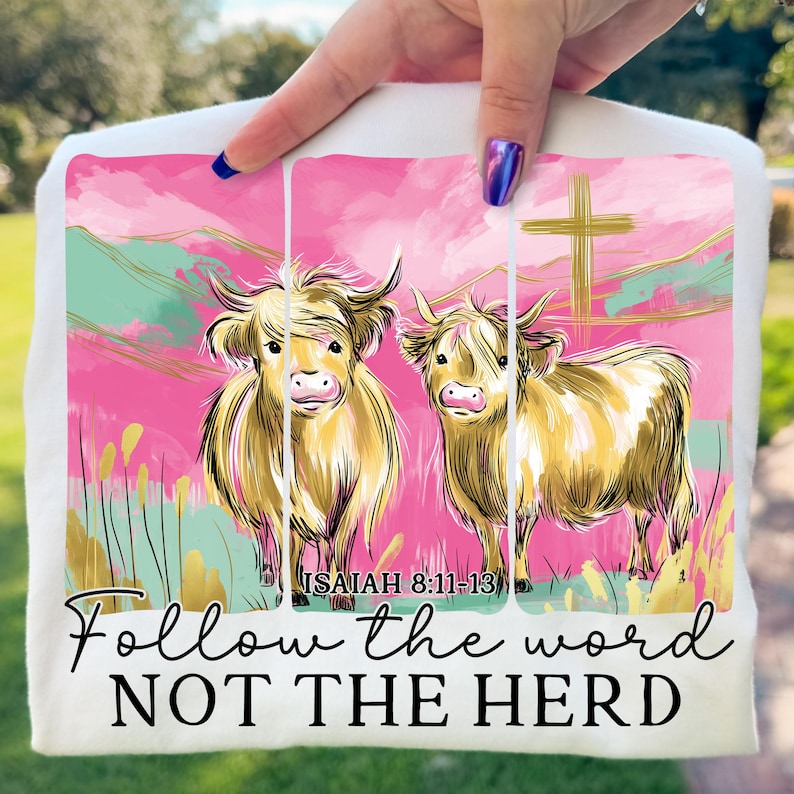 Op de afbeelding: Wit T-shirt met een kleurrijke illustratie van twee Highland-koeien tegen een roze en groene achtergrond, met een gouden kruis en de tekst "Follow the word NOT THE HERD". De tekst "Jesaja 8:11-13" is ook inbegrepen.