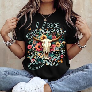 Może przedstawiać: Czarny t-shirt z motywem czaszki byka i napisem "Do No Harm But Take No Bull". Czaszka otoczona jest kolorowymi kwiatami. Koszulka ma okrągły dekolt.