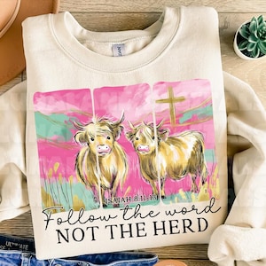 Op de afbeelding: Cr&egrave;mekleurige sweatshirt met een afbeelding van twee Schotse Hooglandrunderen tegen een roze en turquoise achtergrond. Het ontwerp bevat een gouden kruis en de tekst "Follow the word NOT THE HERD" en "Isaiah 8:11-13".