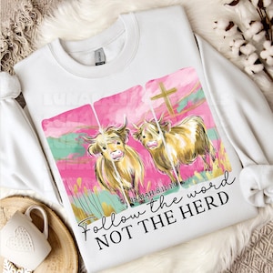 Op de afbeelding: Witte sweatshirt met een aquarelontwerp van twee Schotse Hooglanders tegen een roze en turquoise achtergrond met een gouden kruis. De tekst "Follow the word NOT THE HERD" staat eronder.