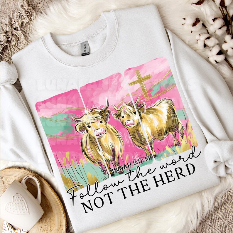 Op de afbeelding: Witte sweatshirt met een aquarelontwerp van twee Schotse Hooglanders tegen een roze en turquoise achtergrond met een gouden kruis. De tekst "Follow the word NOT THE HERD" staat eronder.