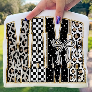 May include: White t-shirt with the word "MAMA" in a bold, decorative font. The letters feature leopard print, a black and white checkerboard pattern, and a starry design. A gray bow graphic is centered on the shirt, with gold glitter accents.