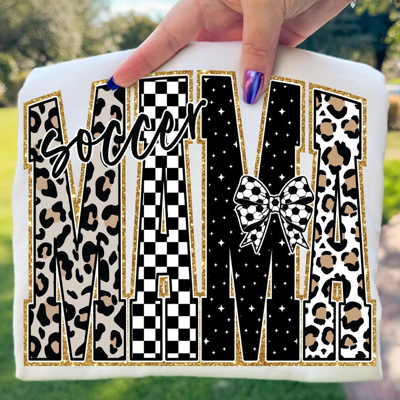 May include: White t-shirt with the words "soccer MAMA" in a gold glitter outline. The letters have various patterns, including leopard print, a black and white checkerboard, and a soccer ball bow.