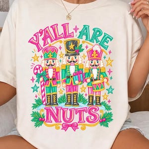 May include: A cream-colored t-shirt featuring a graphic of three nutcracker figures in green, pink, and gold uniforms. The text "Y'ALL ARE NUTS" is printed in pink above and below the figures, surrounded by colorful stars and holly leaves.