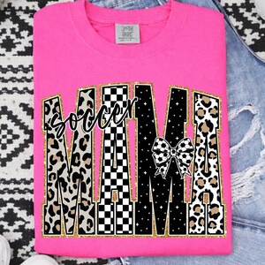 May include: A bright pink t-shirt with the words "Soccer Mama" in a bold, decorative font. The word "Mama" features a mix of leopard print, black and white checkered patterns, and a soccer ball bow. The shirt is folded.