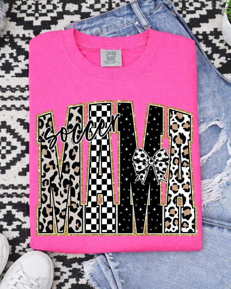 May include: A bright pink t-shirt with the words "Soccer Mama" in a bold, decorative font. The word "Mama" features a mix of leopard print, black and white checkered patterns, and a soccer ball bow. The shirt is folded.