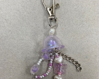 May include: A handmade keychain featuring a clear, iridescent jellyfish charm. The keychain includes a silver clasp, pearl beads, heart-shaped beads, a butterfly charm, and a small chain. The color palette includes shades of purple, white, and silver.