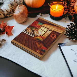 May include: A hardcover journal with a fall-themed illustration. The cover depicts a cozy porch scene with a wreath, flowers, and the words "Hello Fall". The journal is surrounded by autumn decor, including pumpkins, leaves, and a lit candle.