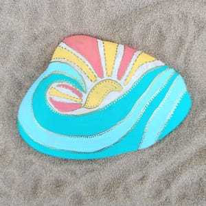 May include: A smooth, irregularly shaped stone painted with a stylized design featuring a yellow and pink sun rising over blue ocean waves. The artwork is outlined with gold-colored dots, creating a decorative pattern across the surface of the rock.