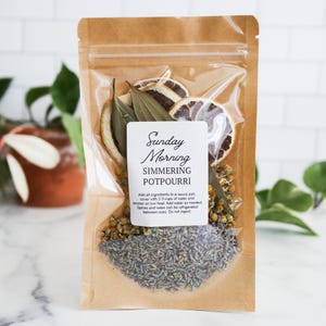 May include: A clear and brown kraft paper pouch containing simmering potpourri with dried orange slices, bay leaves, chamomile flowers, and lavender. A white label reads Sunday Morning SIMMERING POTPOURRI with instructions to add to a sauce pan.