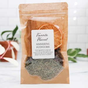 May include: A brown kraft paper pouch containing simmering potpourri with a white label that reads Favorite Flannel, SIMMERING POTPOURRI, and instructions. The contents include a dried orange slice, a cinnamon stick, and dried pine needles.