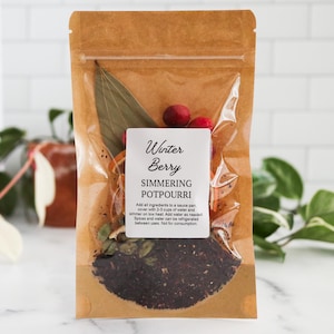 May include: A brown kraft paper pouch containing Winter Berry simmering potpourri. The clear window reveals dried orange slices, bay leaves, red berries, and dark spices. The white label includes instructions: Add all ingredients to a sauce pan, cover with 2-3 cups of water and simmer on low heat. Add water as needed. Spices and water can be refrigerated between uses. Not for consumption.