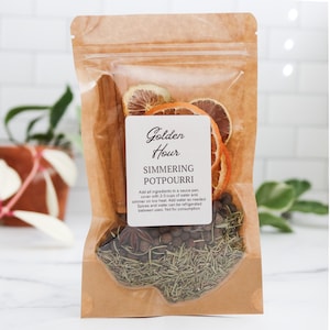 May include: A brown paper pouch containing simmering potpourri with dried orange slices, cinnamon sticks, star anise, and herbs. The white label reads Golden Hour Simmering Potpourri and includes instructions to add 2-3 cups of water and simmer.