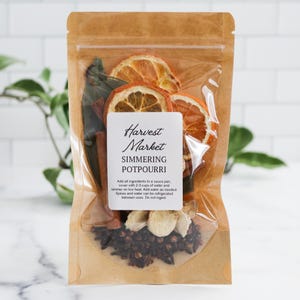 May include: A clear plastic pouch with a brown paper backing containing dried orange slices, cinnamon sticks, bay leaves, and cloves. A white label on the front reads Harvest Market SIMMERING POTPOURRI with instructions to add ingredients to a sauce pan.