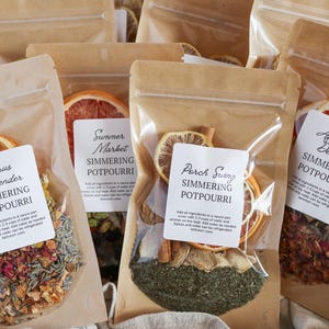 May include: Several clear plastic bags filled with dried botanicals and citrus slices. Each bag has a label with the words "Simmering Potpourri" and a unique scent name like "Citrus Lavender" or "Porch Swing". The bags are sealed and arranged on a light brown surface.