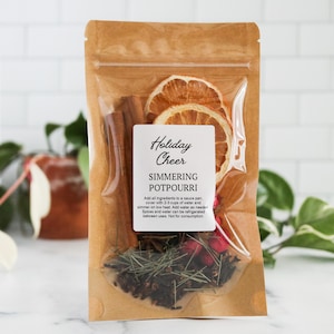 May include: A brown kraft paper pouch containing simmering potpourri with dried orange slices, cinnamon sticks, star anise, and red berries. The white label reads Holiday Cheer SIMMERING POTPOURRI with instructions to add ingredients to a sauce pan.