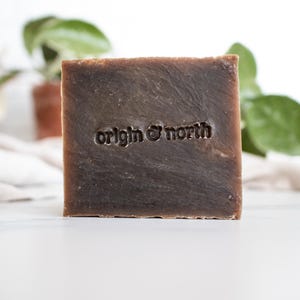 May include: A square bar of brown soap with the embossed text "origin & north". The soap has a textured surface and is set against a white background with green foliage.