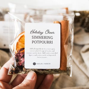 May include: A clear plastic bag filled with simmering potpourri ingredients, including a dried orange slice, cinnamon sticks, and dried flowers. The label reads "Holiday Cheer Simmering Potpourri."