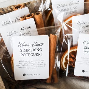 May include: A woven basket filled with clear bags of simmering potpourri. Each bag contains dried orange slices, cinnamon sticks, and other spices. The bags have white labels with the text "Winter Hearth Simmering Potpourri."