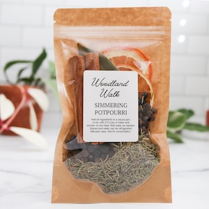May include: A brown kraft paper pouch containing simmering potpourri with visible cinnamon sticks, dried orange slices, dark berries, and green herbs. A white label features the text Woodland Walk SIMMERING POTPOURRI and usage instructions.