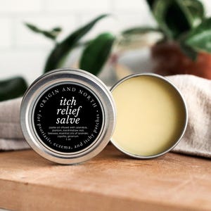 May include: A silver tin of "itch relief salve" with the text "Origin and North" and "for psoriasis, eczema, and itchy patches." The lid is open, revealing a yellow balm. The salve is 1 oz and infused with jojoba oil, calendula, and other ingredients.