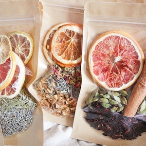 May include: Close-up of several clear plastic bags filled with dried ingredients. The bags contain dried citrus slices, herbs, spices, and tea leaves. The colors are natural, with oranges, reds, greens, and browns. The bags are arranged on a white surface.