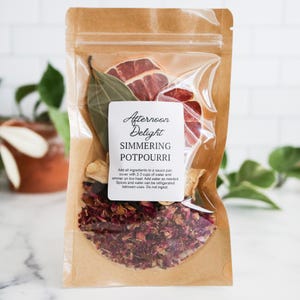 May include: A clear plastic pouch with a brown paper backing containing dried botanicals, including red rose petals, a bay leaf, and dried citrus slices. A white label reads Afternoon Delight SIMMERING POTPOURRI with instructions for use.
