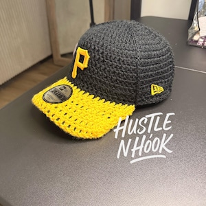 Könnte beinhalten: Handgefertigte Baseballkappe aus schwarzem und gelbem Häkelgarn. Die Kappe hat einen gelben Schirm und ein gelbes "P"-Logo. Der Text "Hustle N Hook" ist in Weiß auf der Oberfläche geschrieben.