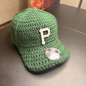Puede incluir: Gorra de béisbol verde tejida a ganchillo con un emblema "P" blanco. La gorra tiene una visera curva y una pegatina "9FORTY ADJUSTABLE". El sombrero está hecho de un material texturizado y tejido.