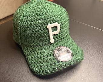 May include: A green crocheted baseball cap with a white "P" emblem on the front. The cap features a curved brim and a "9FORTY ADJUSTABLE" sticker. The hat is made of a textured, knitted material.