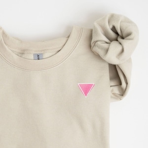 May include: A beige sweatshirt with a pink triangle embroidered on the front.