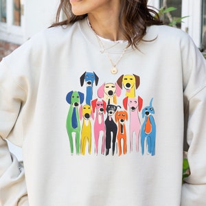 Puede incluir: Una sudadera de color crema con un gráfico colorido de un grupo de perros estilizados. Los perros están representados en varios colores, incluyendo azul, verde, amarillo, rosa y negro. La sudadera tiene mangas largas y cuello redondo.