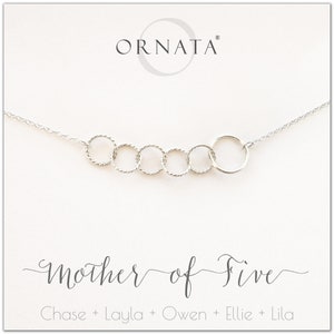 May include: A silver necklace with six interconnected circles. The necklace is on a white card with the text "ORNATA" at the top and "Mother of Five" with five names below. The circles are of varying sizes and textures.