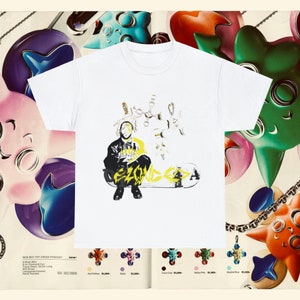 May include: White t-shirt with a graphic of a person wearing a black jacket and jeans, sitting on a skateboard with the word "ZOUK" written in yellow letters. The graphic is surrounded by a chain with multiple charms.