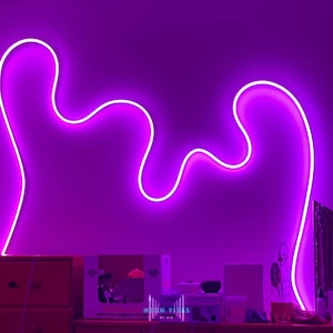 May include: A purple neon light in the shape of a wavy line is mounted on a wall. The light is on and glowing brightly. The wall is a dark purple color.
