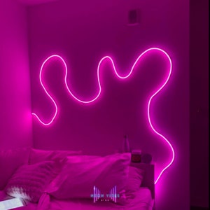Pode incluir: Uma luz de néon rosa em forma de linha ondulada é montada em uma parede acima de uma cama com roupa de cama branca. A luz de néon é etiquetada como "NEON VIBES BY GIO".