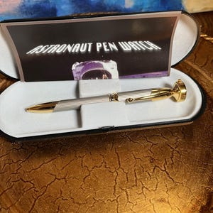 Puede incluir: Un bolígrafo-reloj Astronauta plateado y dorado en un estuche negro. El bolígrafo tiene una punta dorada y un diseño de casco de astronauta dorado en el extremo. El estuche tiene un interior blanco y un exterior negro. El texto "ASTRONAUT PEN WATCH" es visible en una tarjeta dentro.