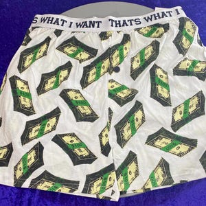 May include: White boxer shorts with a repeated pattern of green and black $100 bills. The waistband has the text "That's What I Want" in black.