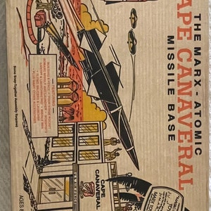 May include: Vintage Marx Toys "Cape Canaveral Missile Base" box art. The illustration features a rocket launch, missile, and a building labeled "U.S. Air Force" with "Cape Canaveral" text. The box is tan with red, black, and yellow graphics.